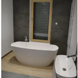 Nowoczesna łazienka z wolnostojącą białą wanną, umywalką nablatową i oknem z drewnianą ramą, wykończona szarą płytką imitującą beton i panelami podłogowymi w odcieniach beżu i szarości.