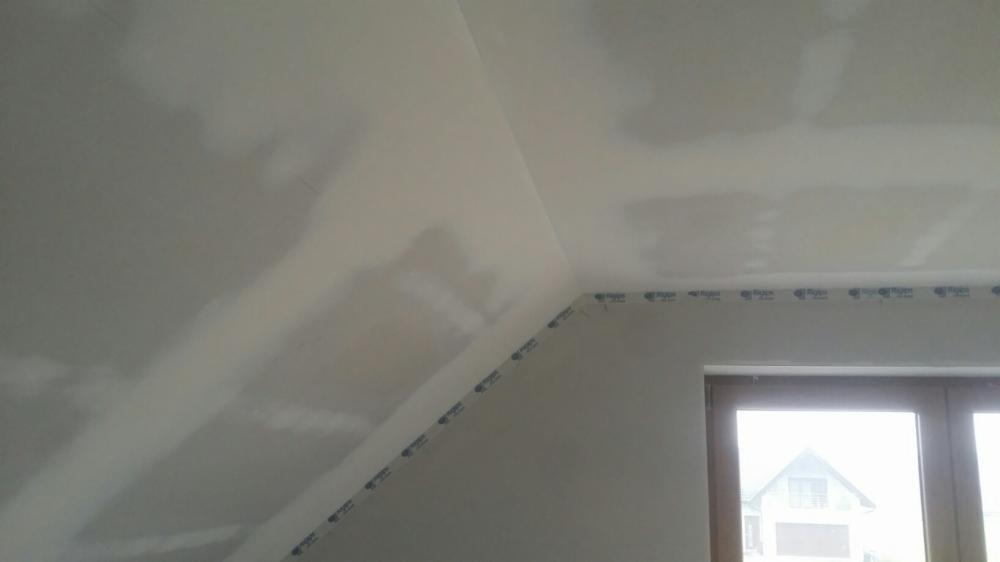 Poddasze z ukończonymi pracami wykończeniowymi płytami kartonowo-gipsowymi, widoczne łączenia płyt przed szpachlowaniem i okno z widokiem na dom.