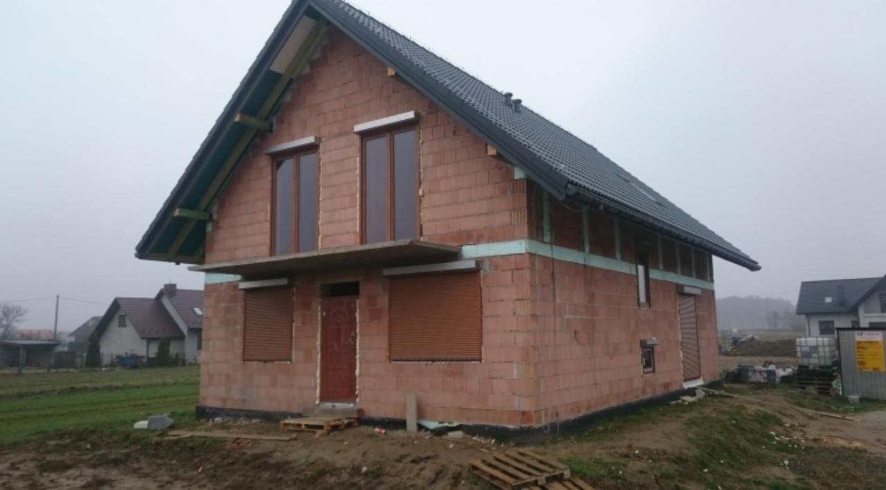 Niedokończony dom jednorodzinny z czerwonej cegły z zamontowanymi oknami i roletami zewnętrznymi, widoczny fragment dachu i ocieplenie, w tle inne domy i pochmurne niebo.