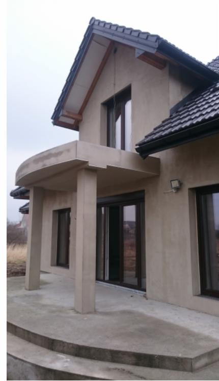 Surowy stan elewacji domu z widocznym zadaszonym wejściem, czarną dachówką i oknami z brązowymi ramami w pochmurny dzień.