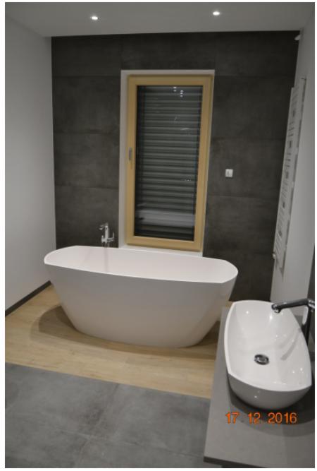 Nowoczesna łazienka z wolnostojącą białą wanną, umywalką nablatową i oknem z drewnianą ramą, wykończona szarą płytką imitującą beton i panelami podłogowymi w odcieniach beżu i szarości.