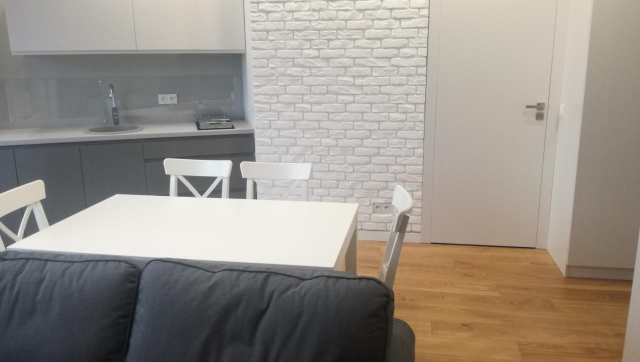 Jasna kuchnia z szarymi szafkami, białą cegłą na ścianie i drewnianą podłogą, widziana z perspektywy salonu z fragmentem szarej sofy.