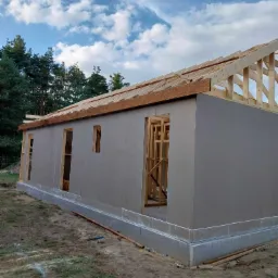 Budowa domu w stanie surowym otwartym: szkielet dachu, ściany z płyt OSB z otworami na okna i drzwi, fundament betonowy, teren budowy z narzędziami.