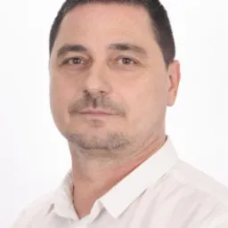 Portret mężczyzny w białej koszuli na jasnym tle, z krótkimi, ciemnymi włosami i delikatnym zarostem. Widoczne zmarszczki mimiczne wokół oczu.