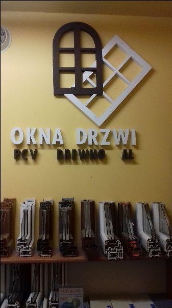 Ekspozycja profili okiennych i drzwiowych w punkcie sprzedaży, z drewnianą i białą ramką okienną na ścianie nad napisem 'OKNA DRZWI'.