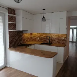 Nowoczesna kuchnia z białymi, lakierowanymi frontami, drewnianym blatem i ceglaną ścianą nad blatem, oświetlona dwoma czarnymi lampami wiszącymi.