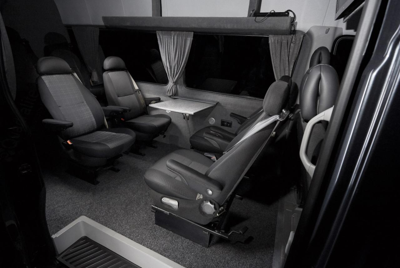 Wnętrze luksusowo wyposażonego busa do przewozu osób z regulowanymi fotelami, rozkładanym stolikiem i zasłonami w oknach.