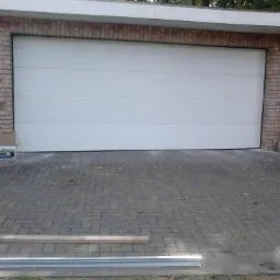 Zamknięta, biała brama garażowa segmentowa z widocznymi elementami konstrukcyjnymi na brukowanym podjeździe, z ceglaną elewacją garażu.