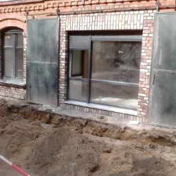 Okna w murze z czerwonej cegły, jedno z roletą zewnętrzną częściowo opuszczoną, obok metalowe drzwi przesuwne na szynie, wykop ziemny przed budynkiem.