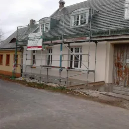 Dom w trakcie renowacji elewacji, widoczne rusztowania i banner firmy budowlanej, drzwi garażowe z widocznymi śladami użytkowania.