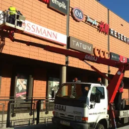 Montaż lub wymiana szyldu Rossmann na elewacji budynku centrum handlowego z użyciem podnośnika koszowego, widoczny pracownik w kamizelce odblaskowej na platformie roboczej.