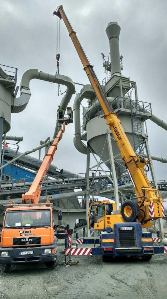 Montaż rurociągu przemysłowego z użyciem żółtego dźwigu samochodowego i pomarańczowego podnośnika koszowego; pracownik w zielonej kamizelce na wysokości łączy elementy, w tle widoczna konstrukcja...