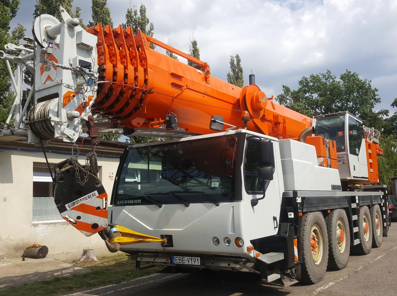 Pomarańczowo-biały dźwig samochodowy Liebherr z wysuniętym teleskopowym ramieniem, zaparkowany na tle budynku i drzew.