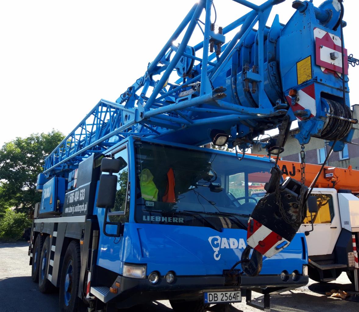 Niebieski dźwig samochodowy Liebherr z wysięgnikiem teleskopowym, widoczne oznaczenia firmy CADA i numer telefonu, zaparkowany na placu budowy.