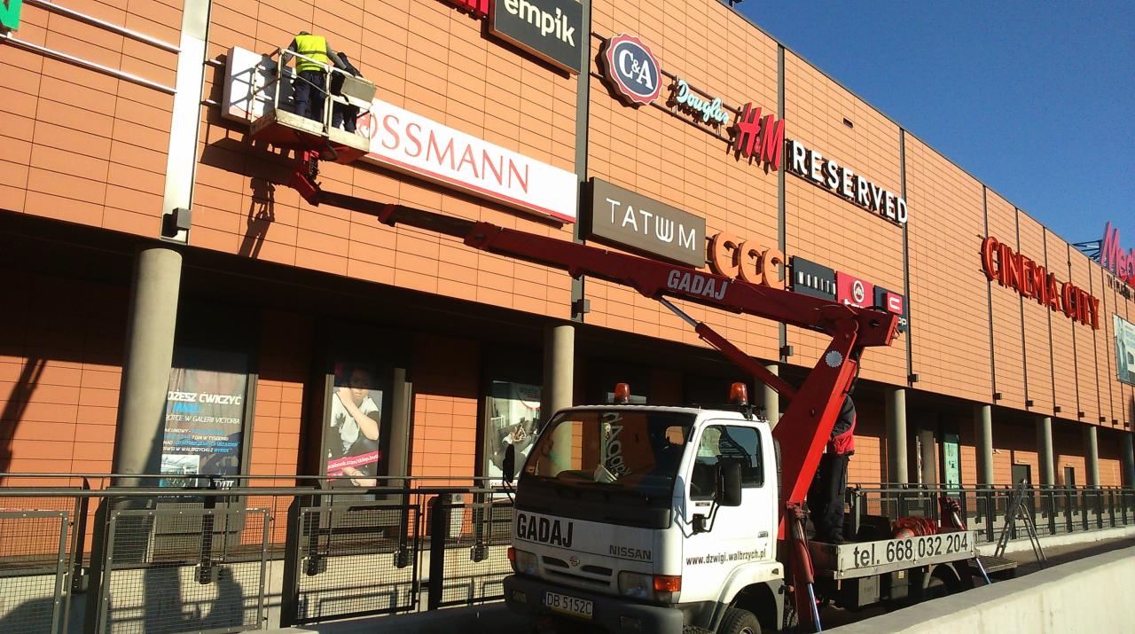 Montaż lub wymiana szyldu Rossmann na elewacji budynku centrum handlowego z użyciem podnośnika koszowego, widoczny pracownik w kamizelce odblaskowej na platformie roboczej.