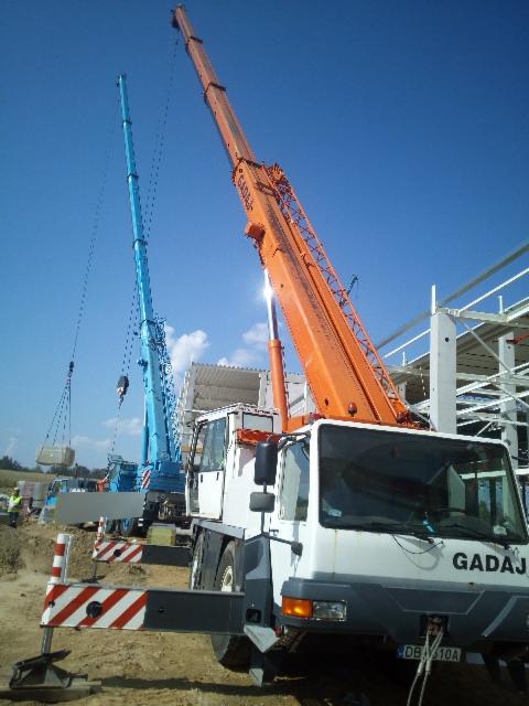 Dwa dźwigi na placu budowy: pomarańczowy dźwig samochodowy z wysięgnikiem pod kątem i niebieski dźwig podnosi betonowy element konstrukcyjny.