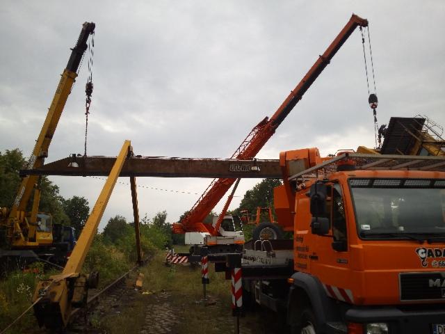 Dwa dźwigi, żółty i pomarańczowy, podnoszą stalową belkę nad torami kolejowymi na tle pochmurnego nieba i zieleni. Widoczna kabina pomarańczowego dźwigu z napisem 'UDZWIG'.
