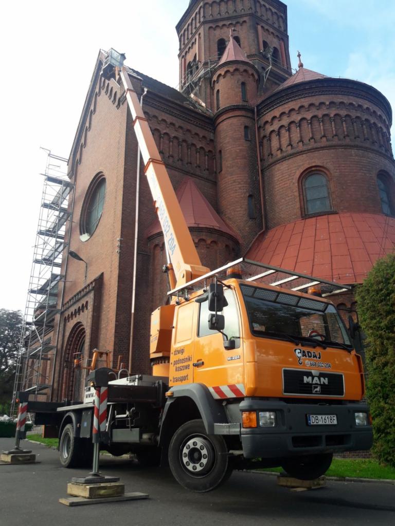Pomarańczowy podnośnik koszowy MAN na drewnianych podkładkach, z wysięgnikiem sięgającym ceglanej fasady kościoła z rusztowaniami, na tle błękitnego nieba.