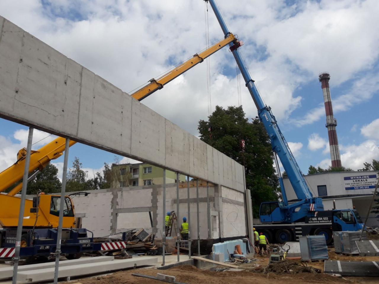 Montaż betonowego prefabrykatu na budowie z użyciem dwóch dźwigów: żółtego i niebieskiego; w tle komin i budynek mieszkalny.