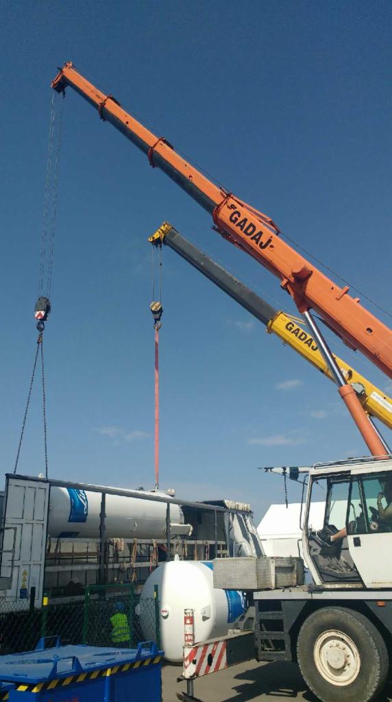 Dwa żurawie teleskopowe firmy GADAJ podnoszą biały zbiornik z niebieskimi oznaczeniami z naczepy ciężarówki na tle błękitnego nieba.