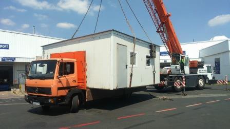 Pomarańczowy dźwig samochodowy podnosi biały kontener biurowy na tle jasnego nieba i budynków przemysłowych.