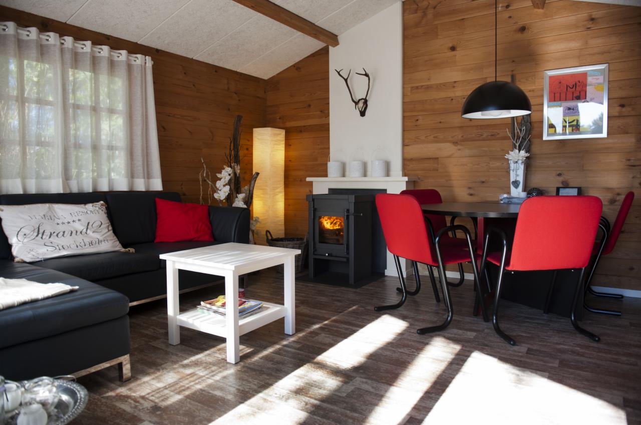 Wnętrze pokoju z drewnianymi ścianami, czarnym piecem na drewno z płomieniami widocznymi przez szybkę, czarną sofą z czerwoną poduszką i białym stolikiem kawowym, oraz okrągłym stołem z czerwonymi...