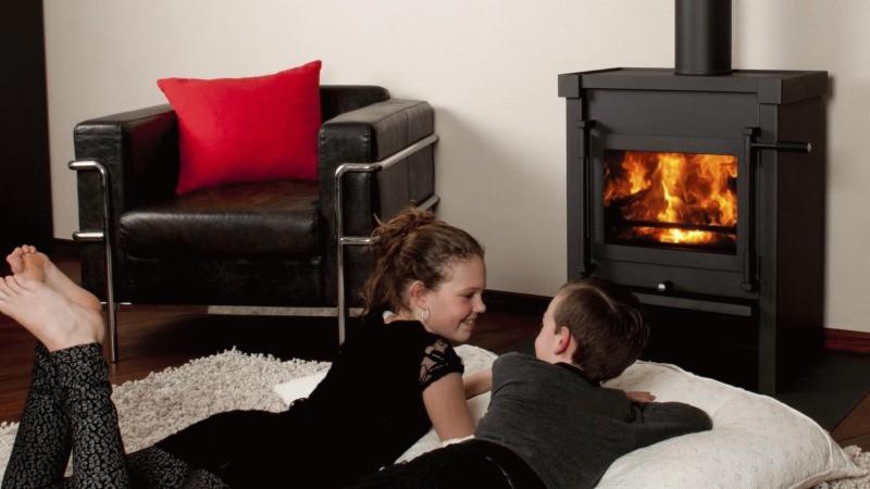 Dzieci leżące na podłodze przed czarnym piecem na drewno, z widocznym płomieniem, w nowoczesnym wnętrzu z czarnym skórzanym fotelem i czerwoną poduszką.