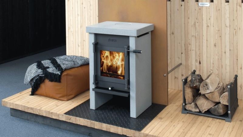 Szary piec na drewno z płomieniami w palenisku, ustawiony na drewnianym podeście z metalową kratką, obok kosz z drewnem i pufa z narzutą w tle.