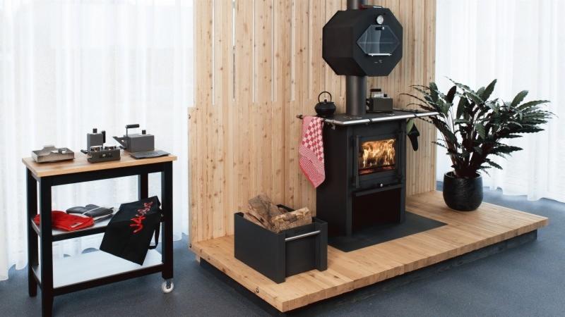 Nowoczesny piec na drewno o geometrycznym kształcie, z paleniskiem w trakcie użytkowania, ustawiony na drewnianym podeście, obok skrzynia z drewnem i roślina doniczkowa, w tle drewniana ściana...