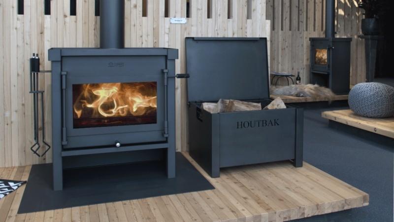 Żeliwny piec na drewno z płomieniami w palenisku, obok otwarty pojemnik na drewno z napisem 'HOUTBAK', drewniana podłoga i ściana z desek.