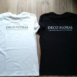 Dwie koszulki, biała i czarna, leżące obok siebie na drewnianej podłodze, z identycznym nadrukiem 'DECO FLORAL Studio Dekoracji Imprez i Pracownia Florystyczna'.