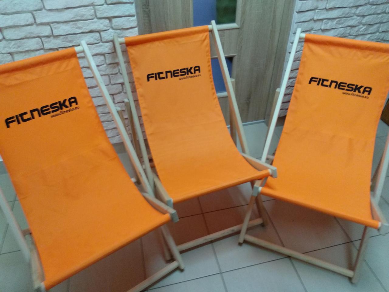 Trzy pomarańczowe leżaki z drewnianymi ramami, z nadrukowanym logo firmy 'FICNESKA' w czarnym kolorze, ustawione obok siebie na podłodze. Tło stanowi jasna ściana z cegły.