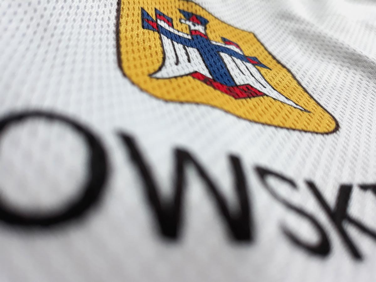 Zbliżenie na białą koszulkę z haftowanym logo orła w kolorach białym, niebieskim i czerwonym na żółtym tle, z widocznym fragmentem czarnego napisu poniżej.