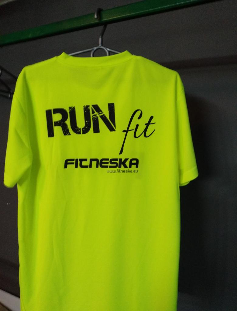 Jaskrawożółta koszulka sportowa z czarnym napisem 'RUN fit' i logo 'FITNESKA' zawieszona na wieszaku na tle szarej ściany.