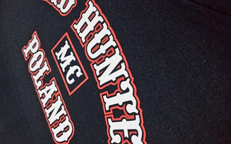 Fragment czarnej koszulki z białym i czerwonym nadrukiem 'HUNTER MC POLAND', widoczne z bliska pod kątem.