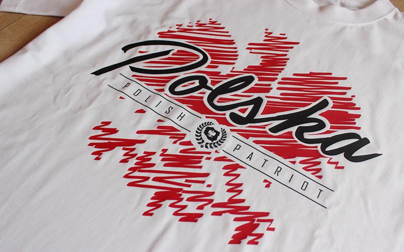 Biała koszulka z czerwono-czarnym nadrukiem konturu Polski i napisem 'Polska Polish Patriot' na drewnianym tle.