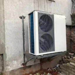 Biała jednostka zewnętrzna pompy ciepła zamontowana na metalowym wsporniku przy szorstkiej, jasnej ścianie budynku, widoczne dwa wentylatory, w tle okno i fragment muru z cegły, gruz budowlany u...