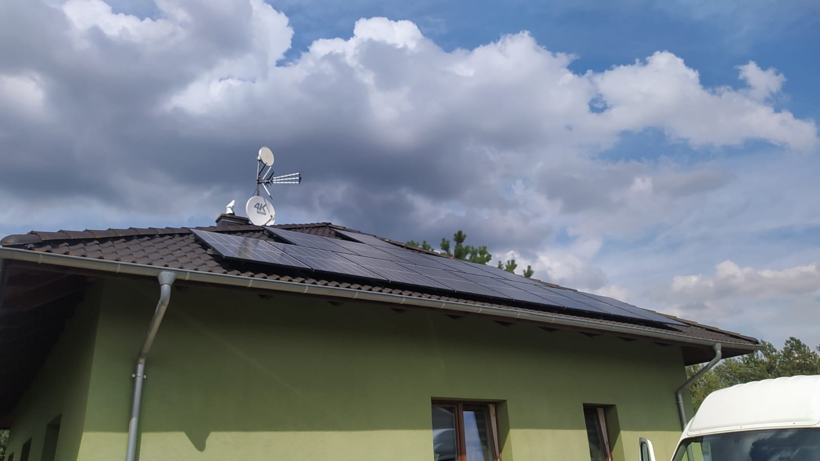 Panele słoneczne na brązowym dachu domu z zieloną elewacją, obok antena satelitarna i fragment białego samochodu dostawczego, na tle pochmurnego nieba.