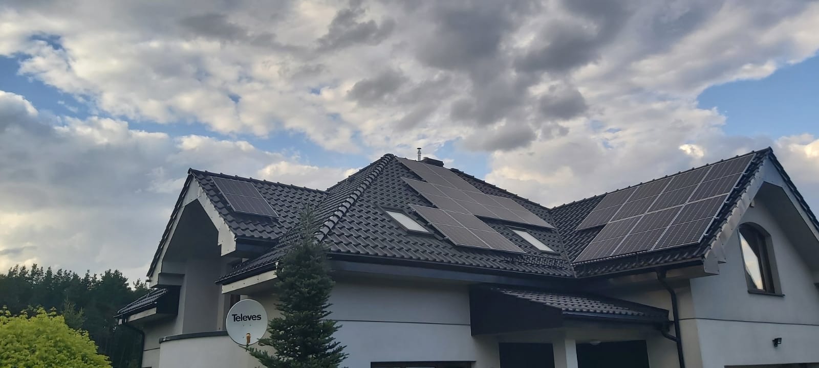 Panele słoneczne na dachu domu jednorodzinnego z ciemną dachówką, widoczne okna dachowe i antena satelitarna.