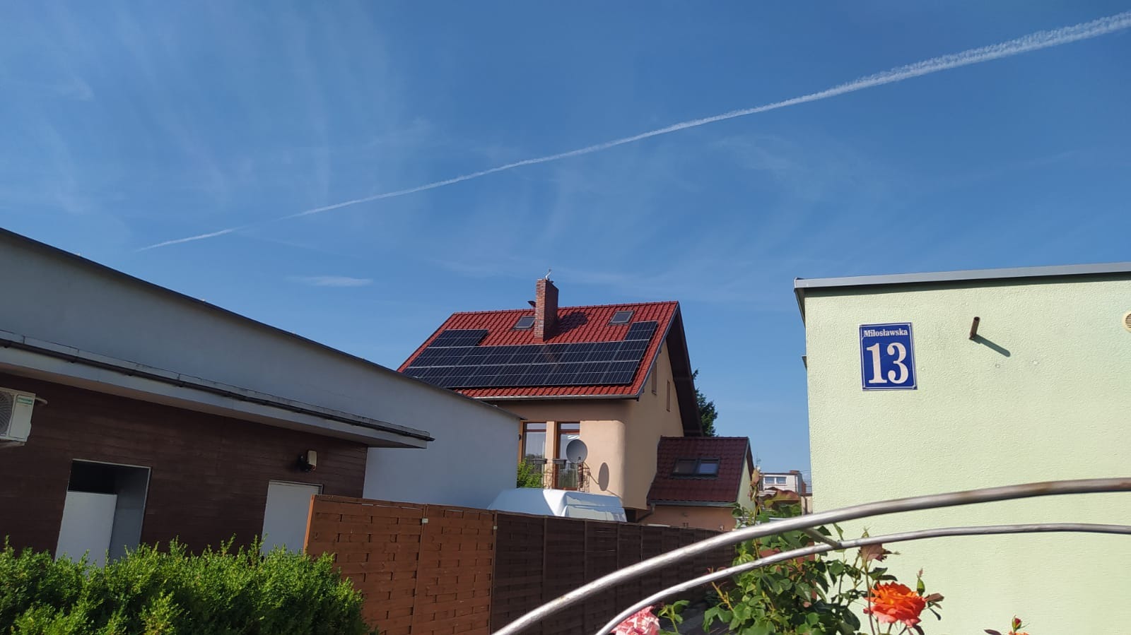 Instalacja paneli słonecznych na dachu domu z czerwoną dachówką w Bolesławicach, widok z poziomu gruntu na tle błękitnego nieba z kondensacyjną smugą samolotu.