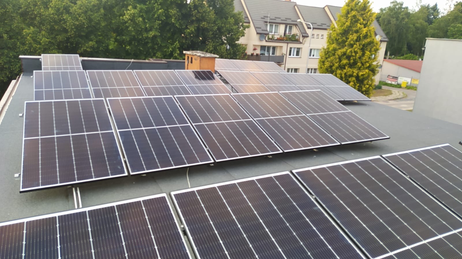 Panele fotowoltaiczne zamontowane na płaskim dachu budynku mieszkalnego, widoczne kominy wentylacyjne i drzewa w tle.