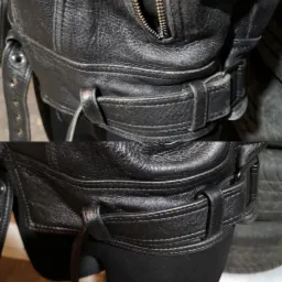 Fragment czarnej, skórzanej kurtki motocyklowej z widocznym paskiem i metalową klamrą, ukazujący detale skóry i szwy.