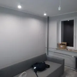 Świeżo pomalowane, szare ściany w pokoju z szarą sofą, widoczne punktowe oświetlenie sufitowe i prowizoryczna lampa wisząca, karton na parapecie.