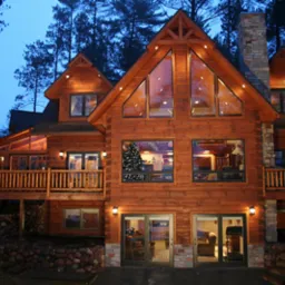 Dwupiętrowy dom z bali w stylu amerykańskim z dużymi oknami, kamiennym kominem i oświetleniem zewnętrznym, otoczony drzewami o zmierzchu.