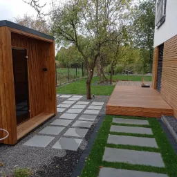 Nowoczesny ogród z drewnianą sauną, tarasem i ścieżką z betonowych płyt, otoczony zielenią i kamieniami.
