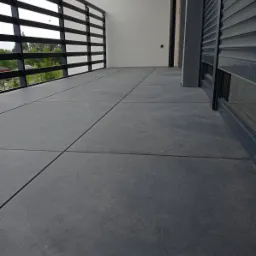 Nowoczesny taras z szarych płyt betonowych, czarna balustrada z poziomych prętów, częściowo zasłonięte rolety okienne w tle.