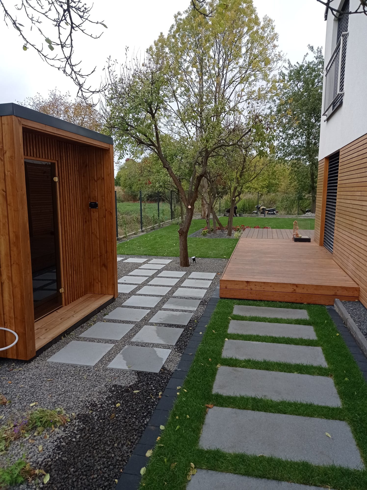 Nowoczesny ogród z drewnianą sauną, tarasem i ścieżką z betonowych płyt, otoczony zielenią i kamieniami.