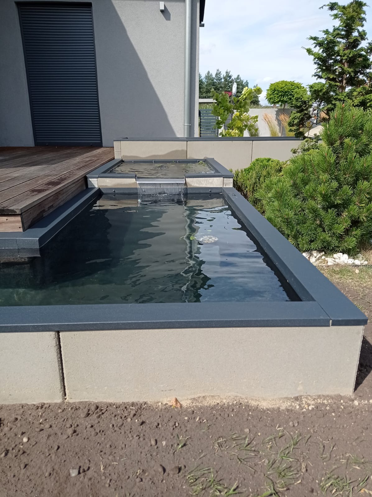 Nowoczesny, kaskadowy zbiornik wodny z szarego betonu i ciemnoszarej obudowy, z małym wodospadem, sąsiadujący z drewnianym tarasem i zielenią ogrodu.