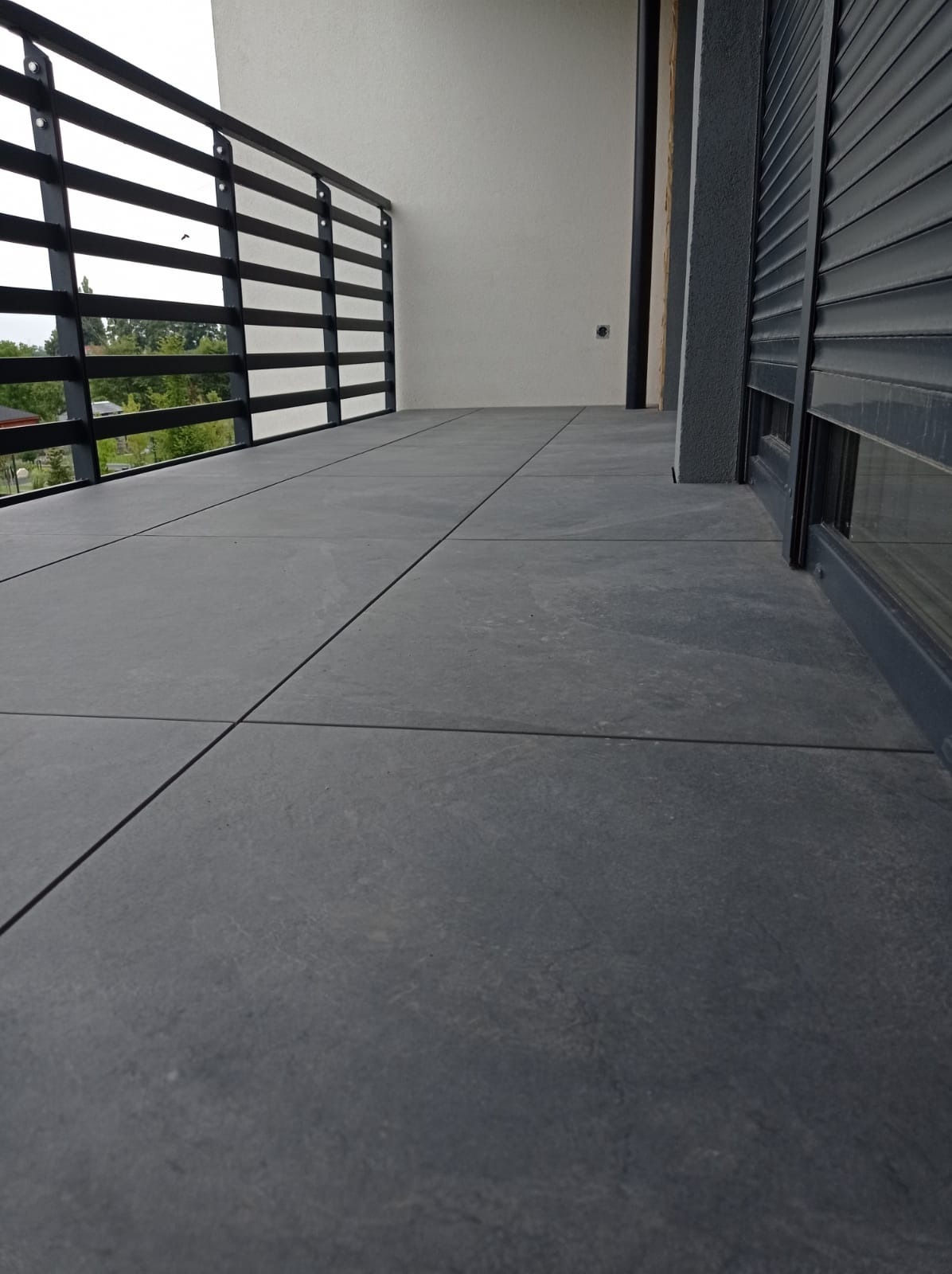 Nowoczesny taras z szarych płyt betonowych, czarna balustrada z poziomych prętów, częściowo zasłonięte rolety okienne w tle.