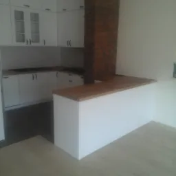 Nowoczesna kuchnia z białymi szafkami, drewnianym blatem i dekoracyjnym kominem z cegły, widok na wyspę kuchenną i fragment podłogi